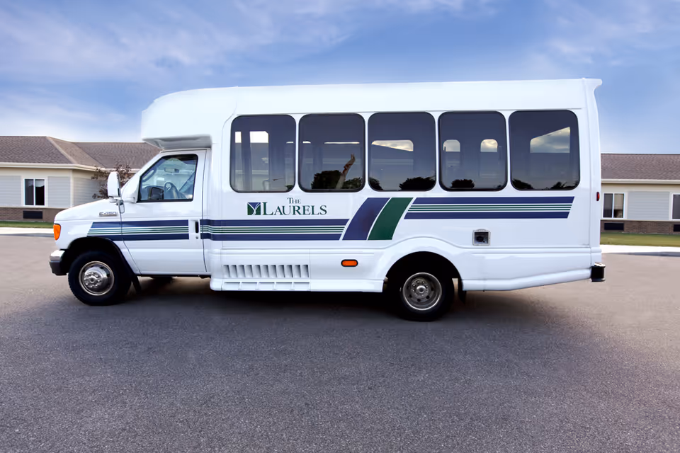 A white shuttle bus with blue and green stripes parked on a paved area in front of a single-story building. The bus has the logo and text 'The Laurels' on its side. The sky is clear with some light clouds.