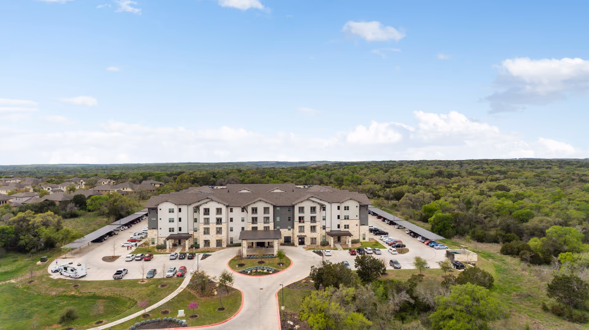 Aerial view of a multi-story senior living building with a circular driveway, parking areas, and surrounding woods.
