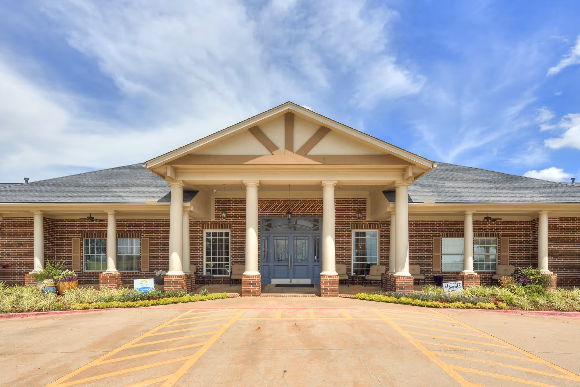 Front exterior view of a single-story brick building with a covered entrance supported by large white columns, a gray double door, and windows on either side. There are chairs on the porch and landscaped greenery along the front. The sky is partly cloudy.