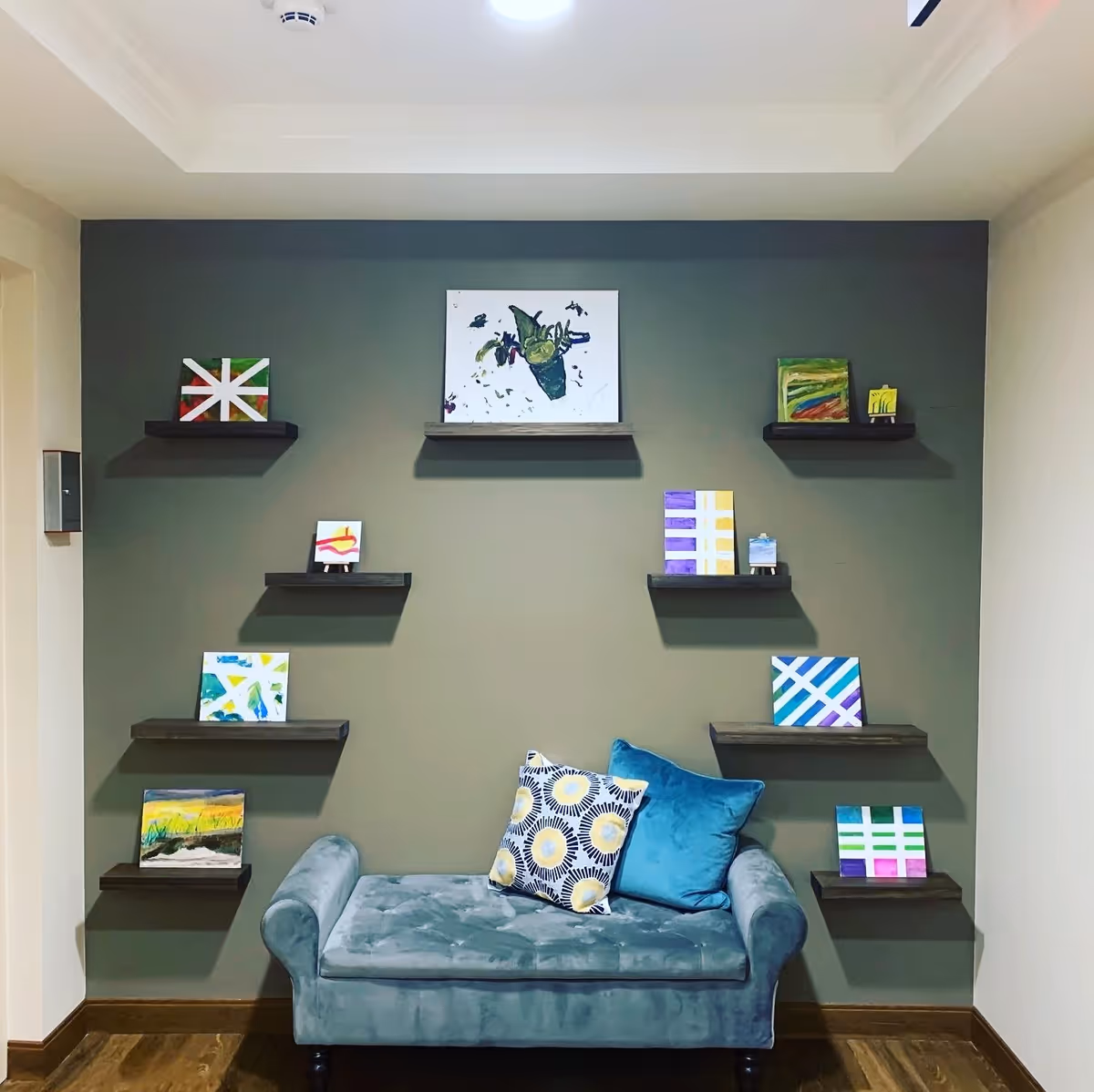 A cozy interior corner featuring a teal velvet loveseat with two decorative pillows, one patterned and one solid teal. The wall behind is painted gray and adorned with nine small floating shelves displaying colorful abstract paintings. The floor is wooden, and the ceiling has a recessed design with a light fixture.