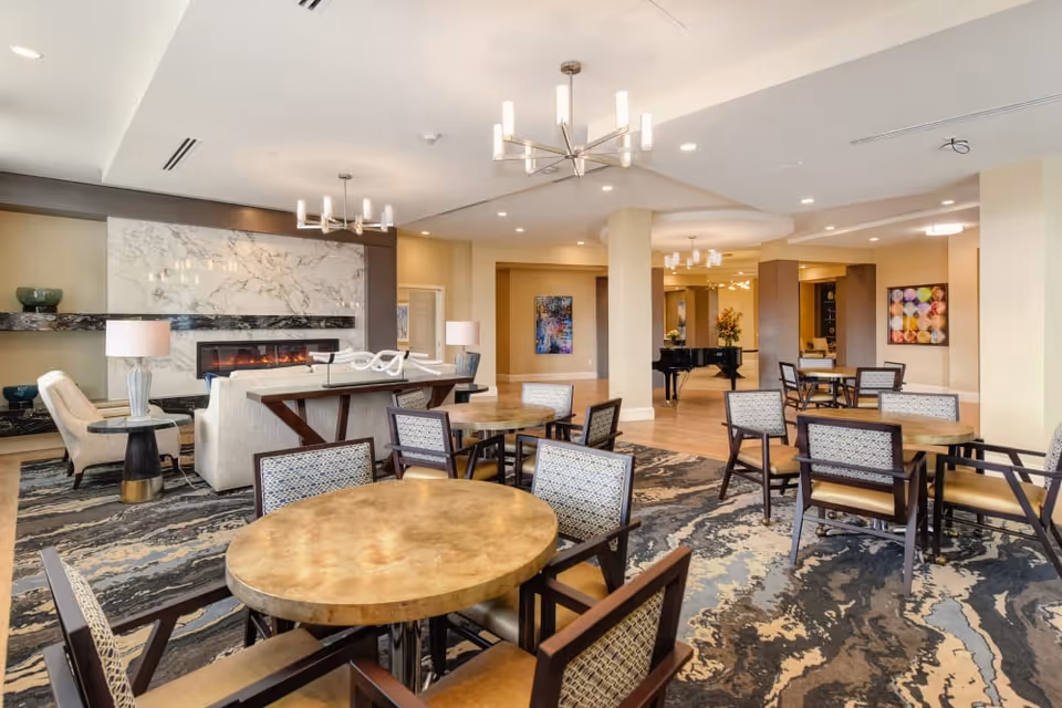 A spacious and well-lit common area in a senior living facility featuring multiple round tables with chairs, a seating area with sofas and armchairs near a modern fireplace with a marble backdrop, decorative lamps, and a grand piano in the background. The room has contemporary chandeliers and colorful artwork on the walls.