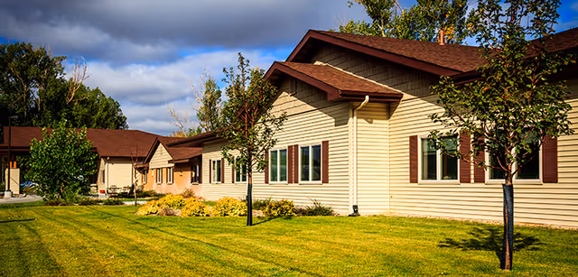 Exterior view of a single-story building with beige siding and brown roof, surrounded by a well-maintained lawn and young trees under a partly cloudy sky.