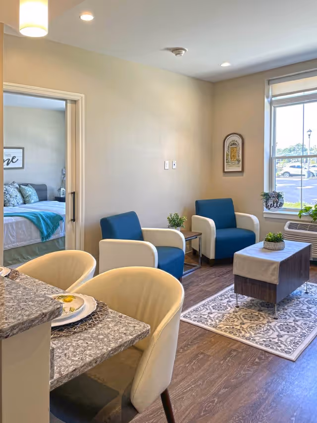 A cozy senior living interior featuring a small living area with two blue and white armchairs, a wooden coffee table with a decorative runner and a small plant, a patterned area rug, and a window with a view of parked cars outside. Adjacent to the living area is a dining counter with two cream-colored chairs and place settings. Through an open doorway, a bedroom with a bed, pillows, and a teal throw blanket is visible.