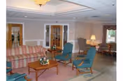 A cozy living room area in a senior living facility featuring a striped sofa, two blue armchairs, a wooden coffee table with a small floral arrangement, a side table with a lamp, and a window with curtains. The room has soft lighting and neutral-colored walls with double doors in the background.