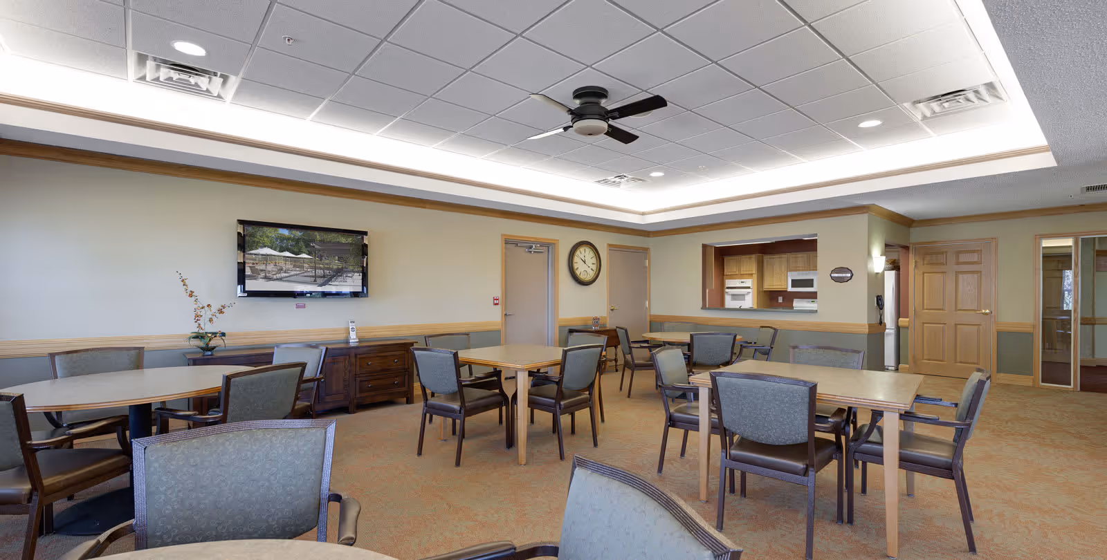 A spacious common area with multiple tables and chairs arranged for group seating. The room features a ceiling fan, a wall-mounted TV displaying an outdoor scene, a clock on the wall, and a pass-through window to a kitchen area with wooden cabinets and appliances. The walls are painted in neutral tones with wood trim, and the carpet is a warm beige color.