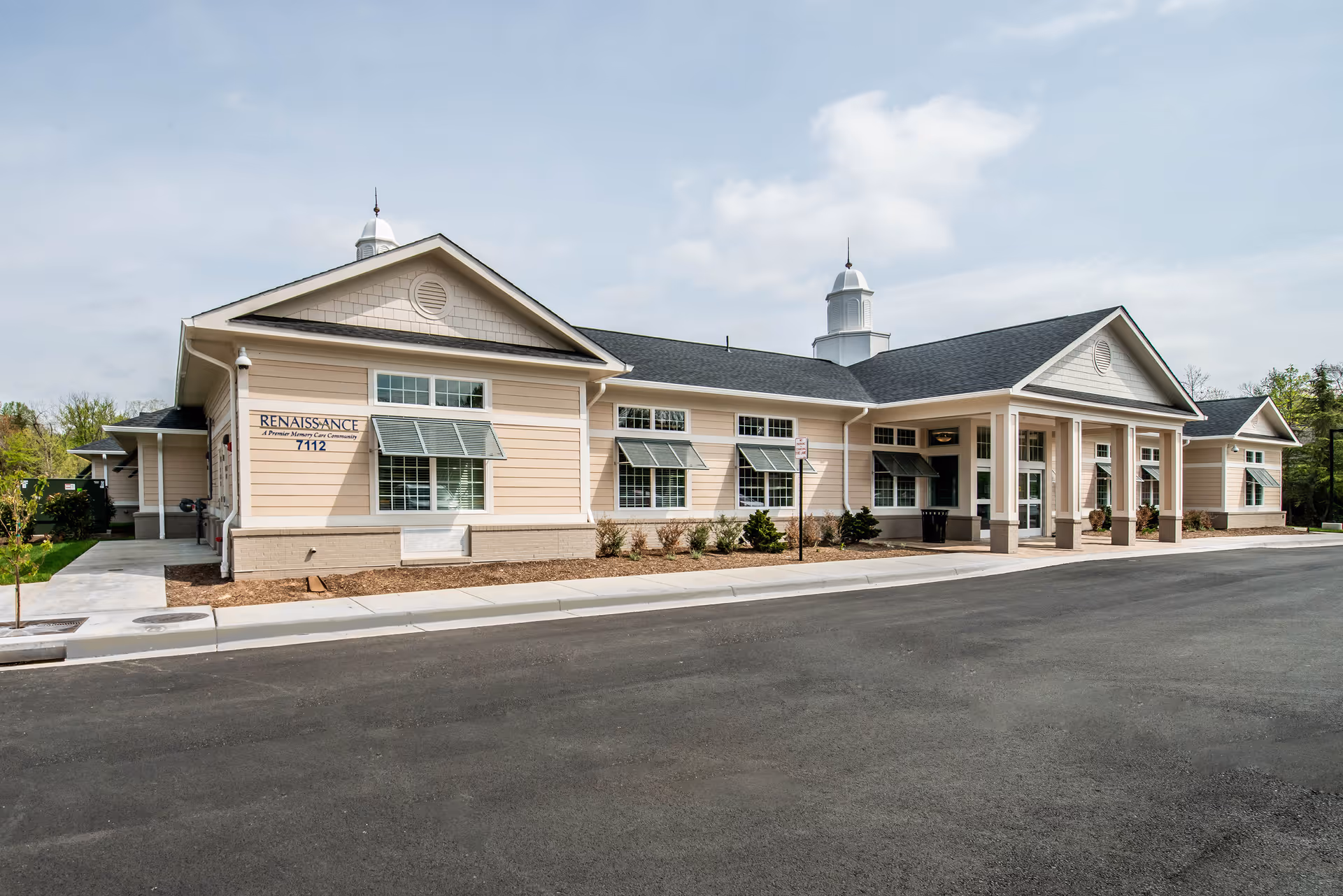 Exterior view of Renaissance of Annandale, a single-story senior living facility with beige siding, multiple windows with green awnings, a covered entrance supported by columns, and a paved driveway in front.