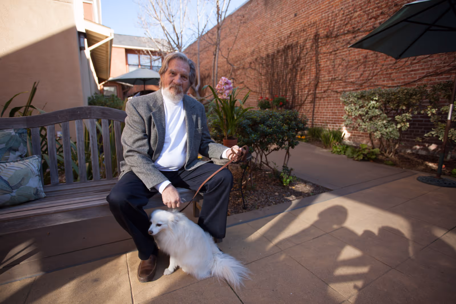 An elderly man with gray hair and a beard sits on a wooden bench in an outdoor courtyard area. He is wearing a gray blazer, white shirt, and dark pants, holding a leash attached to a small white fluffy dog sitting beside him. The courtyard has plants, flowers, and a brick wall in the background with some outdoor umbrellas providing shade.