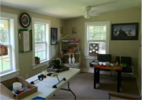A well-lit room with beige walls and carpeted floor featuring a white folding table with various items on it, a small foosball table, a metal shelving unit with supplies, and several windows letting in natural light. The room has framed pictures on the walls and a ceiling fan.
