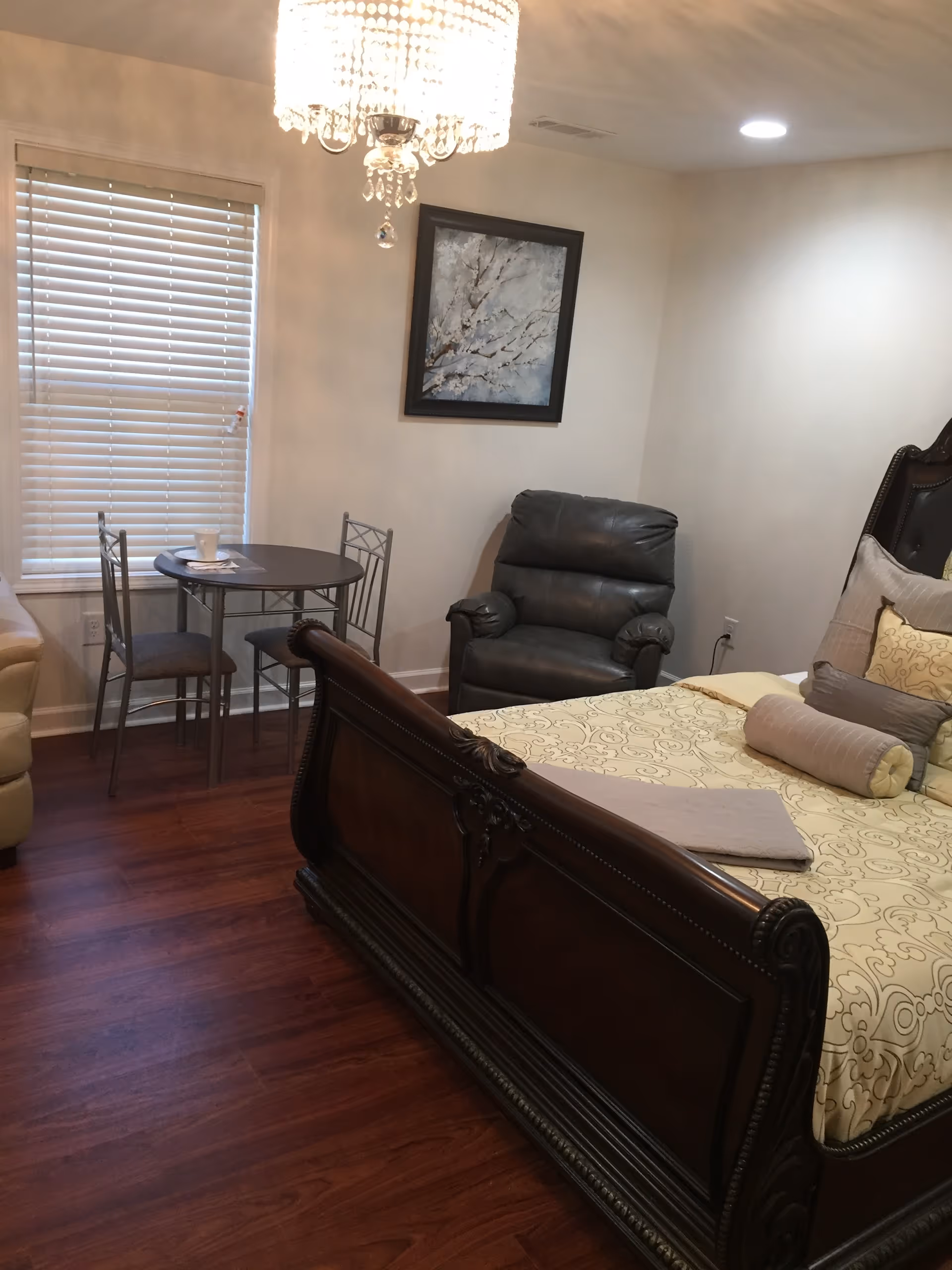 A cozy bedroom with a large wooden bed featuring decorative pillows and a beige patterned bedspread. There is a black leather armchair in the corner, a small round table with two metal chairs near a window with closed blinds, and a framed painting of a tree on the wall. A chandelier hangs from the ceiling, and the floor is a polished dark wood.
