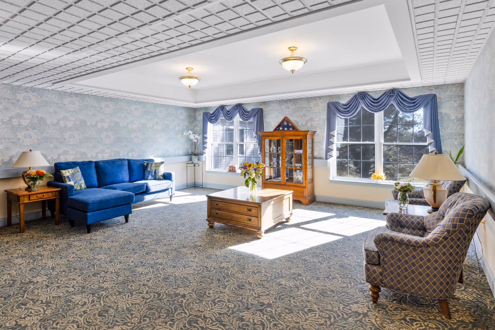 A bright and spacious living room with patterned carpet and wallpaper. The room features a blue upholstered sofa with floral pillows on the left, two patterned armchairs on the right, and a wooden coffee table with a vase of flowers in the center. There are two large windows with blue valance curtains letting in natural light. A wooden display cabinet with a folded American flag is positioned between the windows. Two ceiling light fixtures illuminate the room.