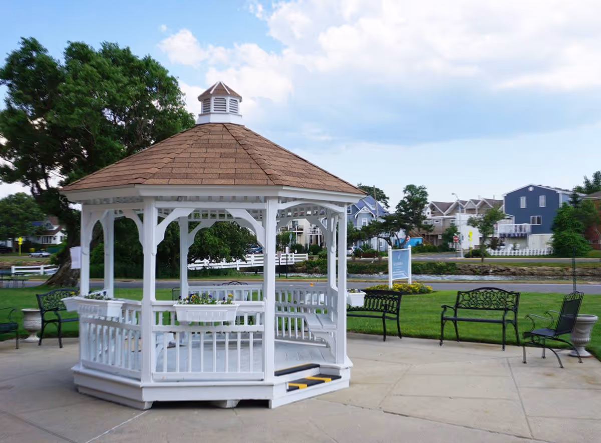 White wooden gazebo with a brown shingled roof situated on a concrete patio surrounded by green grass and black metal benches. In the background, there are trees, a road, and residential houses under a partly cloudy sky.