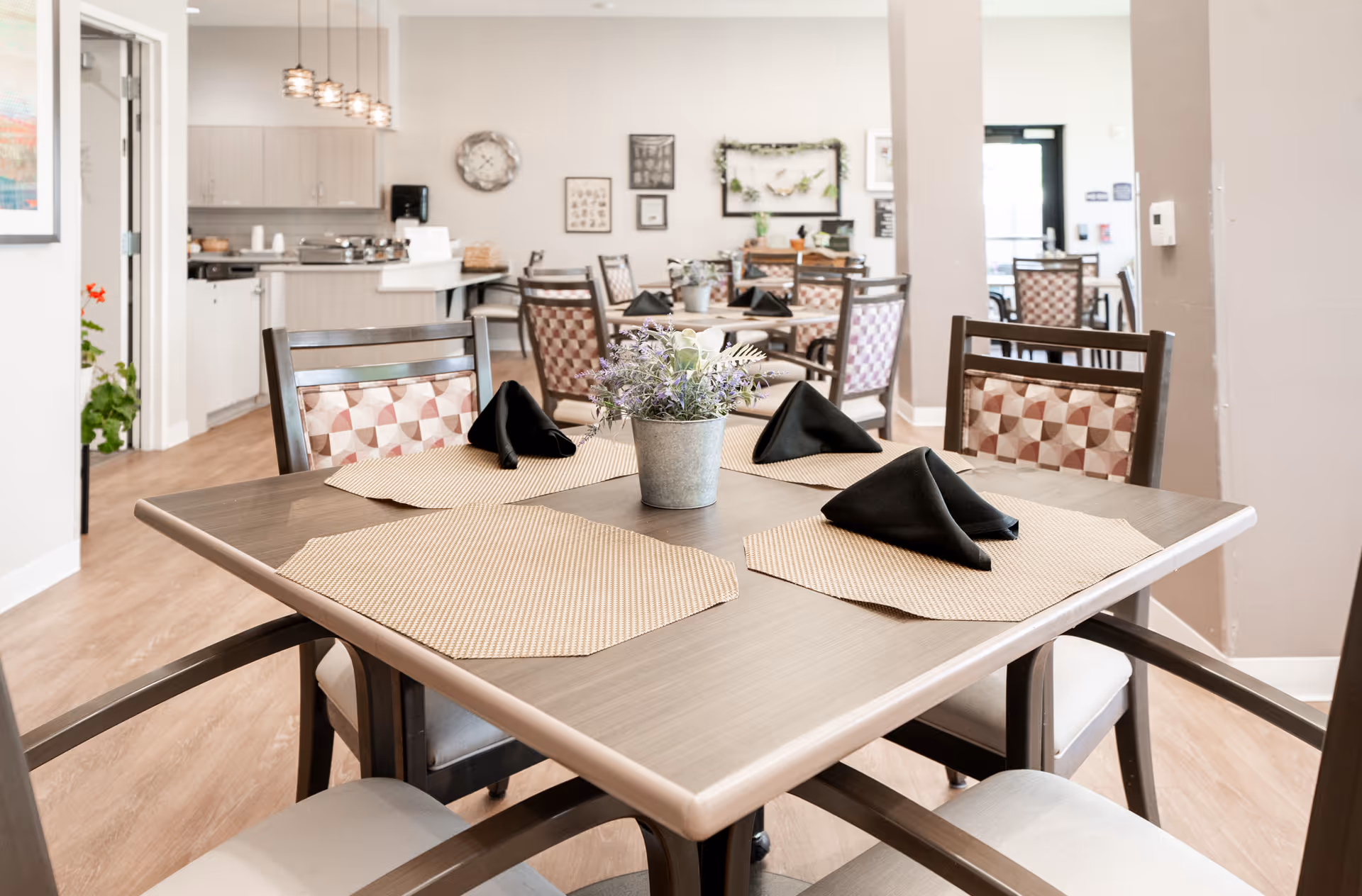 Bright communal dining room with a wooden table set with placemats, folded black napkins, and a small potted plant, with chairs and a kitchenette in the background.