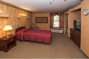 Spacious senior living bedroom with a double bed in a red bedspread, nightstand and lamp, TV on a cabinet, seating, and a window with curtains.