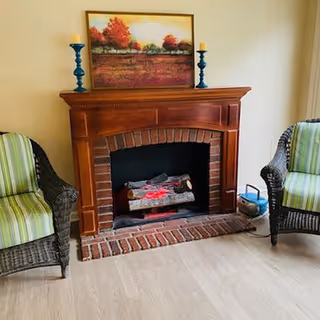 A cozy indoor setting featuring a wooden fireplace with a brick interior and faux logs. On the mantel above the fireplace, there is a framed painting of a landscape with trees and a field, flanked by two candle holders with candles. On either side of the fireplace are wicker armchairs with green and white striped cushions. The floor is light-colored wood, and a small radio or speaker is placed on the floor to the right of the fireplace.