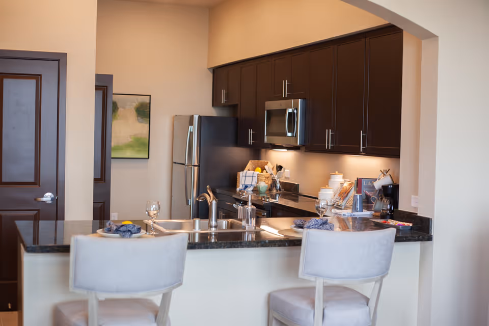 Modern kitchen with dark wood cabinets, stainless steel refrigerator and microwave, granite countertops, and a breakfast bar with two light gray upholstered chairs. The kitchen is well-lit with under-cabinet lighting and has various kitchen items and decor on the counters.