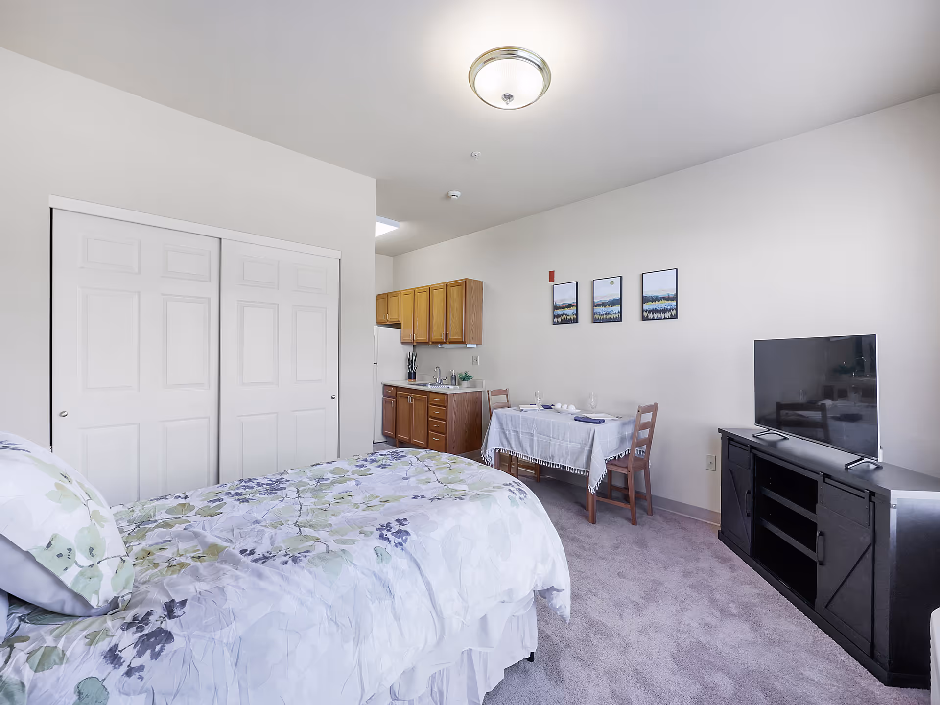 A senior living facility room featuring a bed with floral bedding, a small dining table with two chairs set with glasses and napkins, a flat-screen TV on a black TV stand, a kitchenette with wooden cabinets and a refrigerator, and a closet with white sliding doors. The walls are white and decorated with three framed landscape pictures.