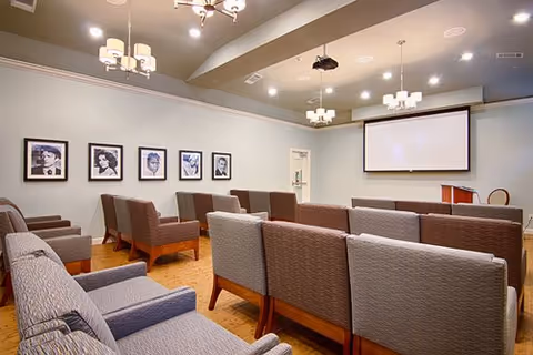 A small auditorium or presentation room with several rows of cushioned chairs facing a large projection screen. The walls are decorated with framed black and white portraits. The ceiling has multiple modern light fixtures and a projector mounted in the center.