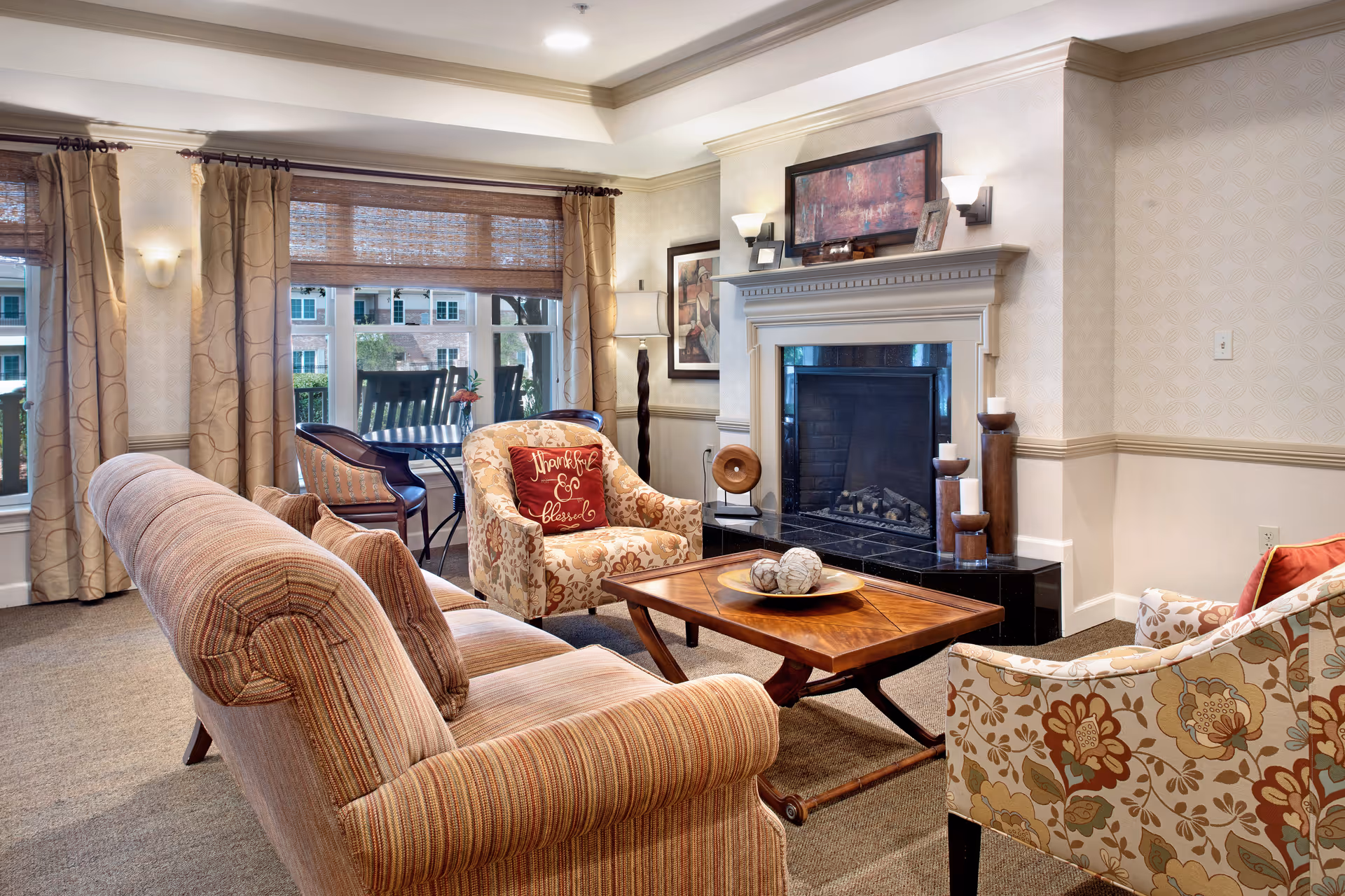 A cozy living room with a striped sofa, two floral armchairs, a wooden coffee table with decorative balls, and a fireplace with candles and framed artwork above it. Large windows with beige curtains and woven blinds let in natural light, and a small table with two chairs is visible near the window.