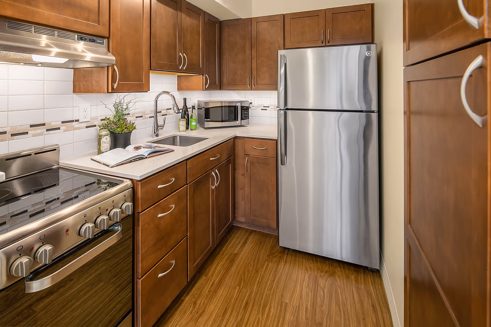 A modern kitchen with wooden cabinets, a stainless steel refrigerator, a stainless steel stove with oven, a microwave, a sink with a faucet, and a countertop with a small plant, two bottles, and an open book. The backsplash is white tile with a decorative strip, and the floor is wood.