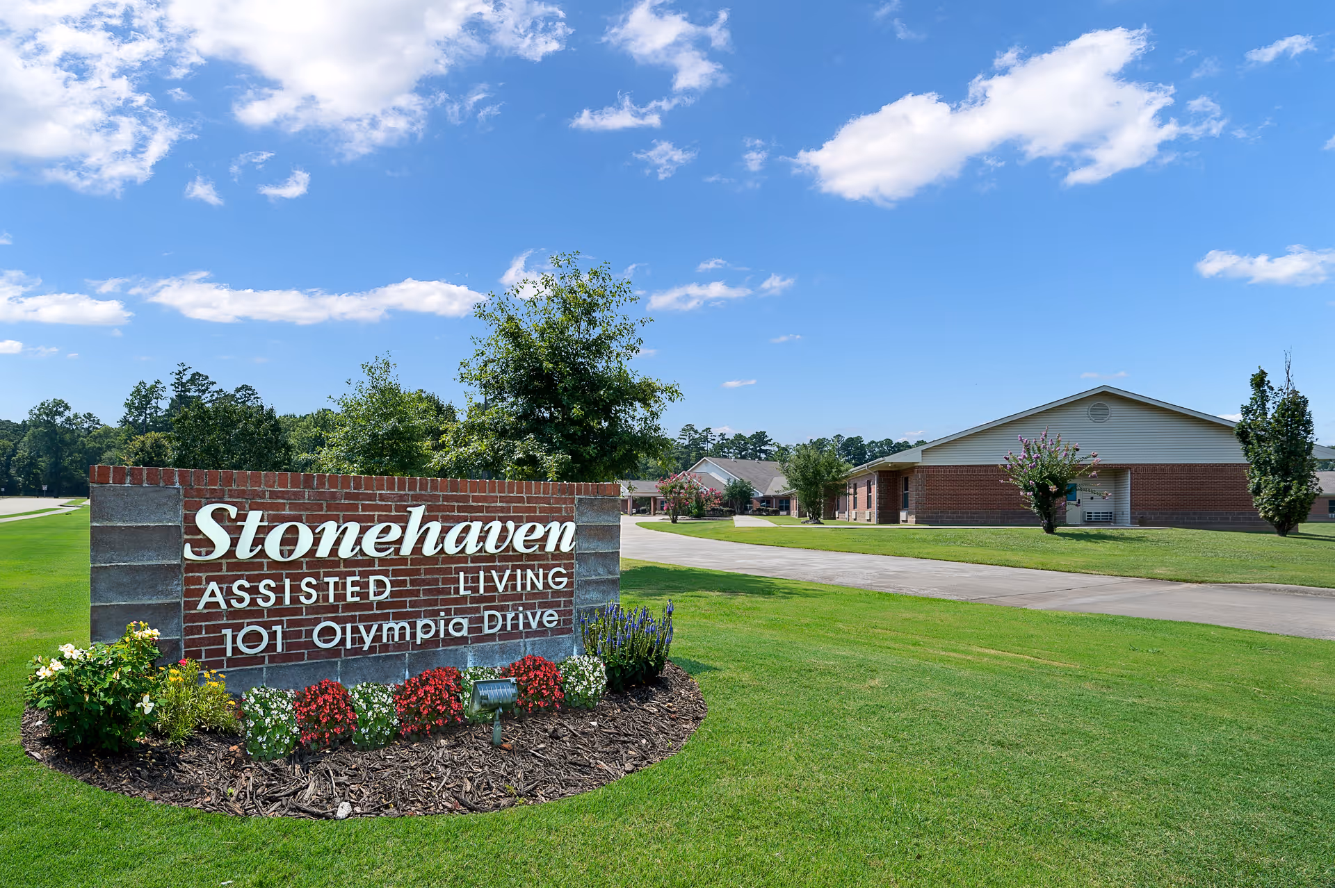 Brick sign for Stonehaven Assisted Living at 101 Olympia Drive surrounded by flowers and green grass, with a clear blue sky and buildings in the background.
