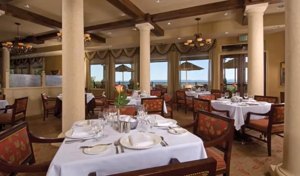 Elegant dining room with tables set in white linens, glassware, floral centerpieces, and columns with windows looking out to umbrellas.