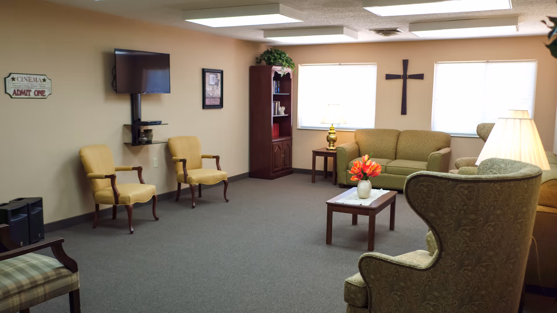 A cozy living room area in a senior living facility with beige walls and carpeted floor. The room features a green sofa, two yellow armchairs, a patterned armchair, a wooden coffee table with a vase of red flowers, a wooden bookshelf, two windows with blinds, a wall-mounted TV, and a cross hanging on the wall between the windows. There are also two table lamps providing warm lighting.