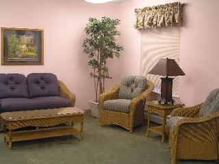 A cozy living room with a wicker sofa and two wicker armchairs with cushions, a wicker coffee table, a side table with a lamp, a tall potted plant, a window with blinds and a valance, and a framed painting on a light pink wall.