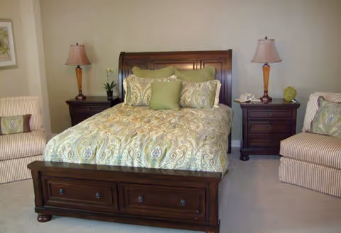 A bedroom with a wooden bed frame featuring a paisley-patterned bedspread in shades of green and beige. The bed is flanked by two dark wooden nightstands, each with a lamp and decorative items. Two striped armchairs with matching pillows are positioned on either side of the bed against light-colored walls.