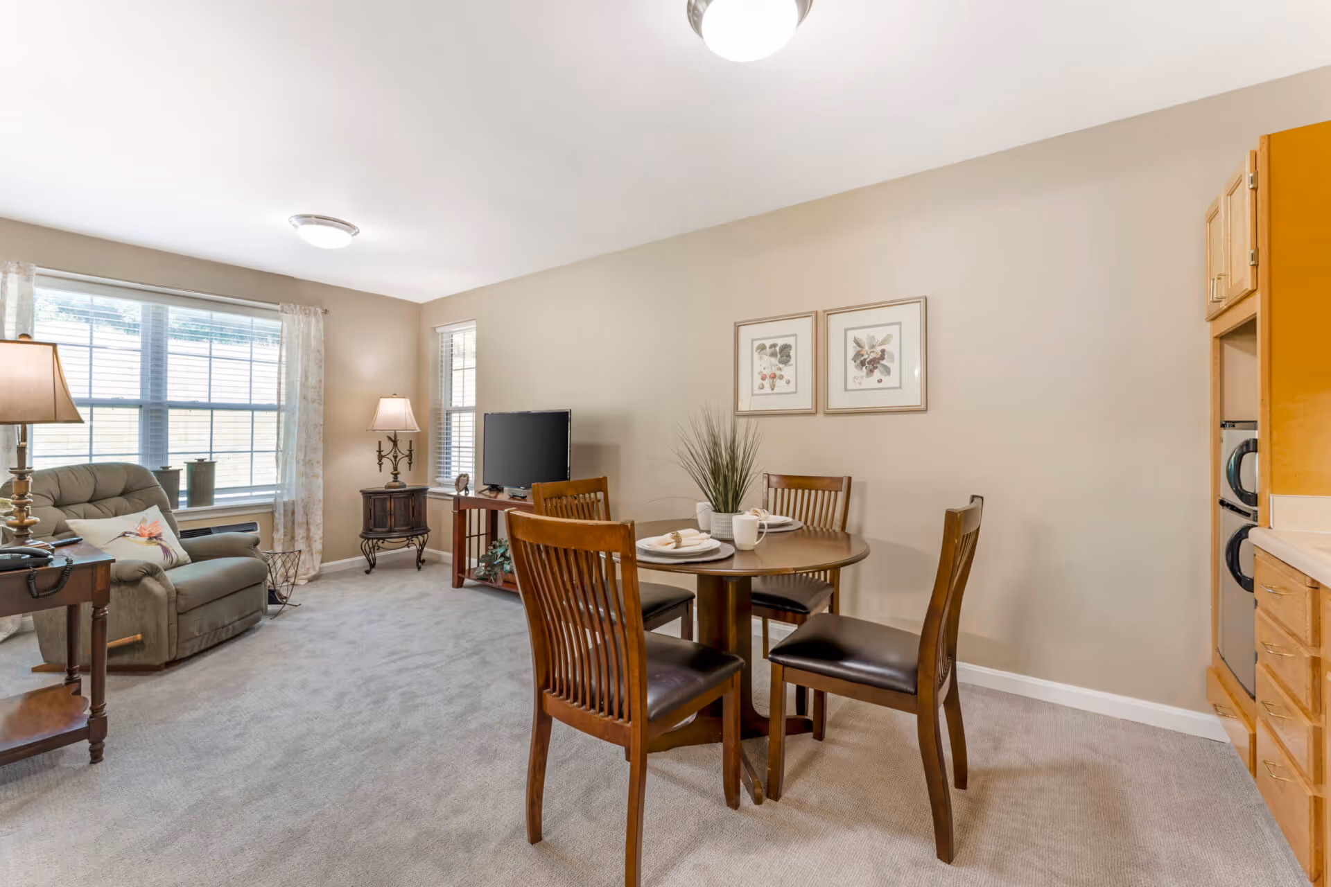 Bright combined living and dining area with a round wooden table and chairs, a recliner by a large window, TV on a small stand, and neutral decor.