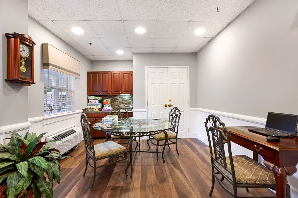 A well-lit room with a round glass table surrounded by four ornate chairs with cushioned seats. On the table, there are several board games and puzzles. To the right, there is a wooden desk with a laptop and mouse on it, accompanied by a matching chair. The room has wood flooring, light gray walls with white trim, a window with blinds and a beige valance, and a wall-mounted wooden clock. In the background, there are wooden cabinets and a double white door.