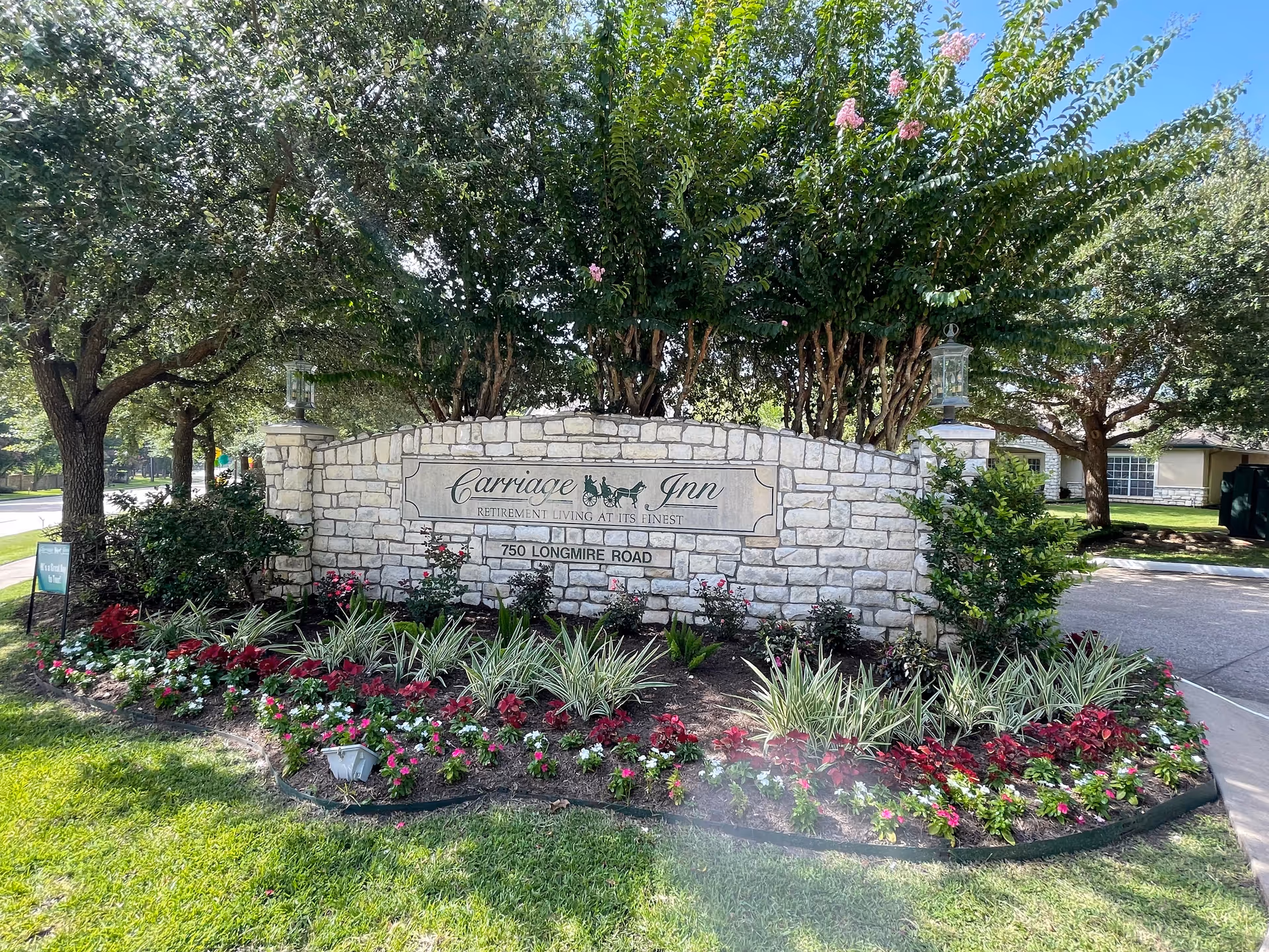 Stone sign for Carriage Inn retirement living surrounded by landscaped flowers and greenery, with trees and a clear blue sky in the background.