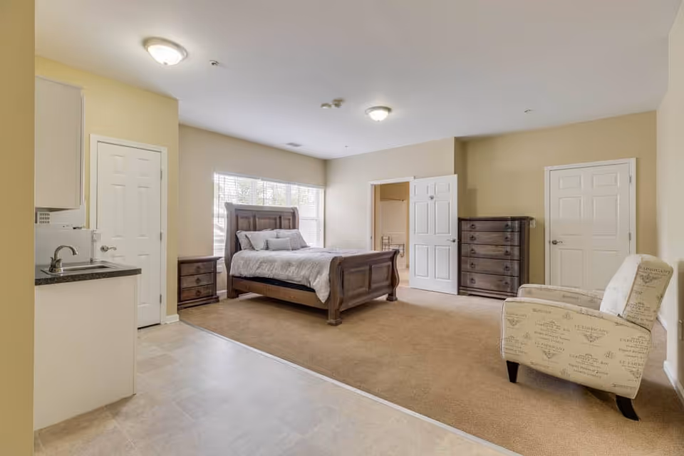 A spacious senior living bedroom with a large wooden bed, two nightstands, a tall dresser, and a comfortable upholstered armchair. The room has beige walls, carpeted flooring, and a small kitchenette area with a sink and countertop on the left side. There are two closed white doors and one open door leading to another room, with a large window behind the bed letting in natural light.