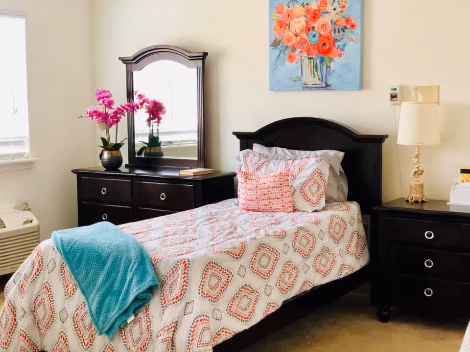 A neatly made single bed with patterned bedding and a turquoise throw blanket in a bright bedroom. The room features dark wood furniture including a dresser with a mirror and a nightstand with a lamp. A colorful floral painting hangs on the wall above the bed, and a potted pink orchid sits on the dresser near a window.