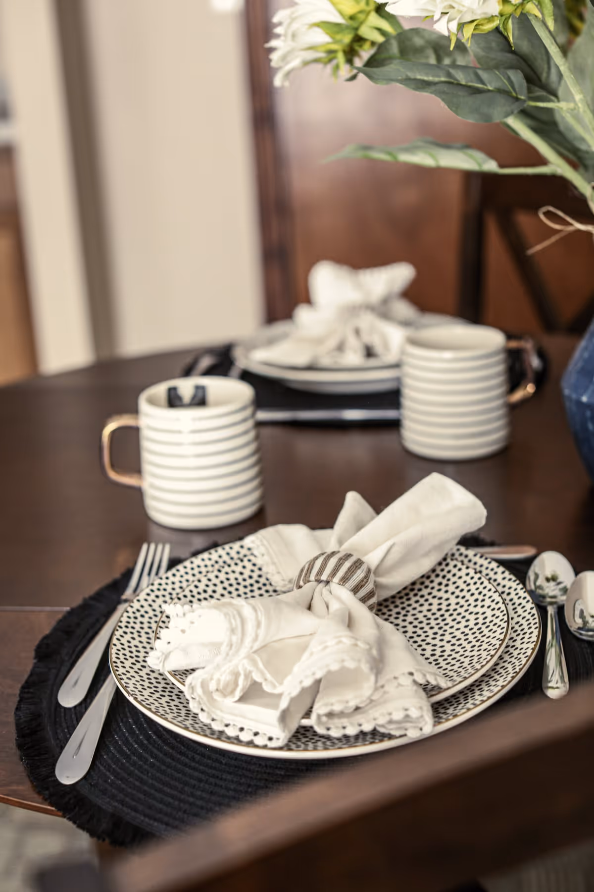 Close-up of a dining table set with patterned plates, white cloth napkins with napkin rings, striped mugs, and silverware on a dark placemat. A vase with white flowers is partially visible on the right side.