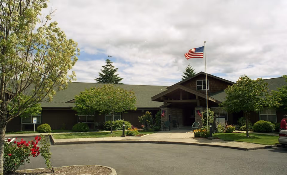 Front entrance of a single-story assisted living building with an American flag, landscaped trees, and a circular driveway.