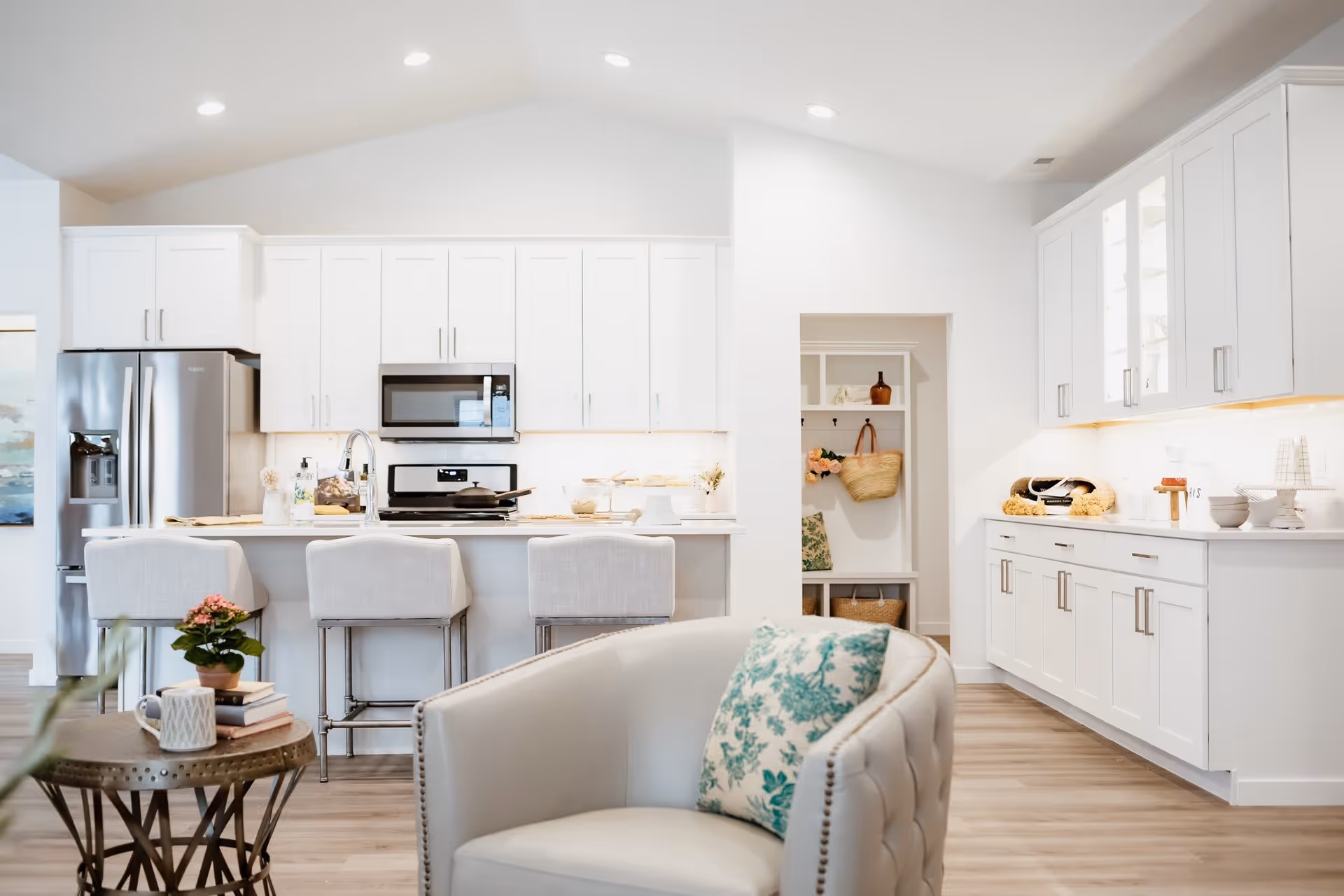 Bright open-concept kitchen with white cabinetry, a central island with three stools, stainless steel appliances, and a seating area in the foreground.