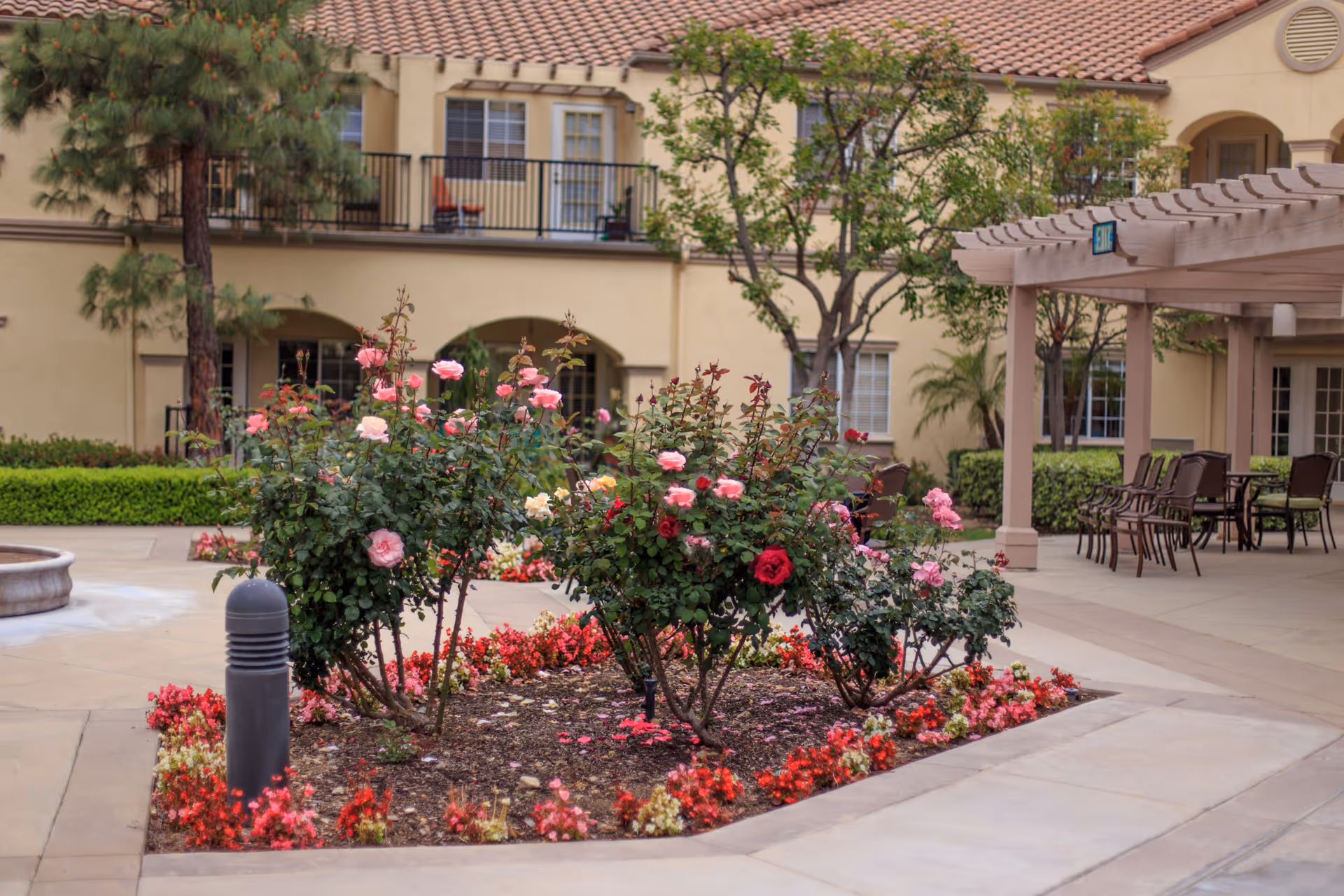 A courtyard garden area with blooming pink and red roses surrounded by a paved walkway. There are outdoor chairs and tables under a pergola to the right, and a beige building with balconies and windows in the background.