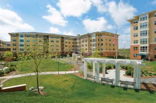 Outdoor view of a senior living facility with multiple connected buildings surrounding a landscaped garden area featuring green lawns, young trees, and a white pergola structure under a partly cloudy sky.