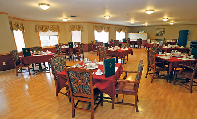 A spacious dining room with multiple tables covered in red tablecloths, each set with cups, saucers, and menus. The room has wooden chairs with patterned upholstery, large windows with valances, and warm lighting from ceiling fixtures.