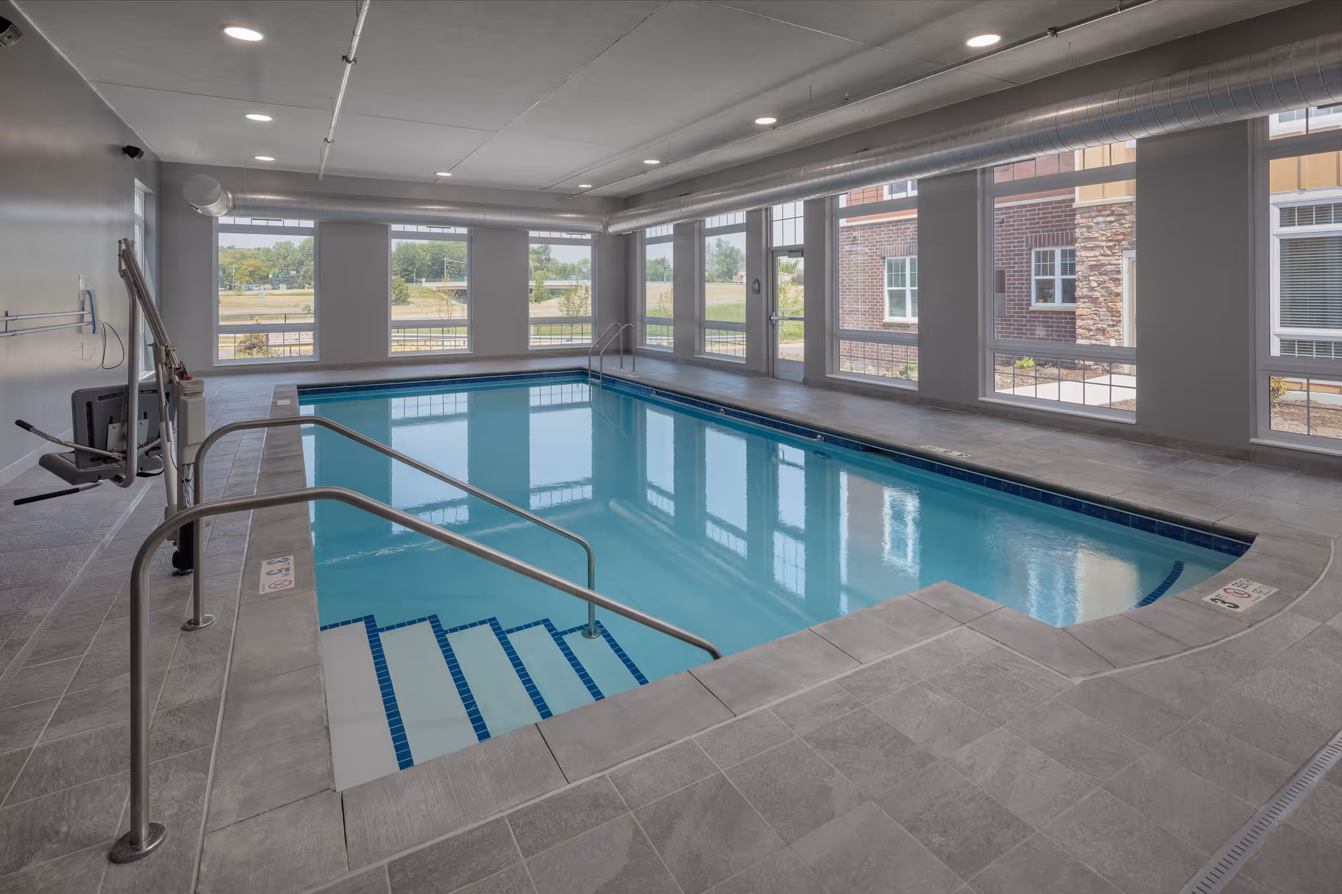 Indoor swimming pool with steps, handrails, a pool lift and large windows overlooking the grounds.