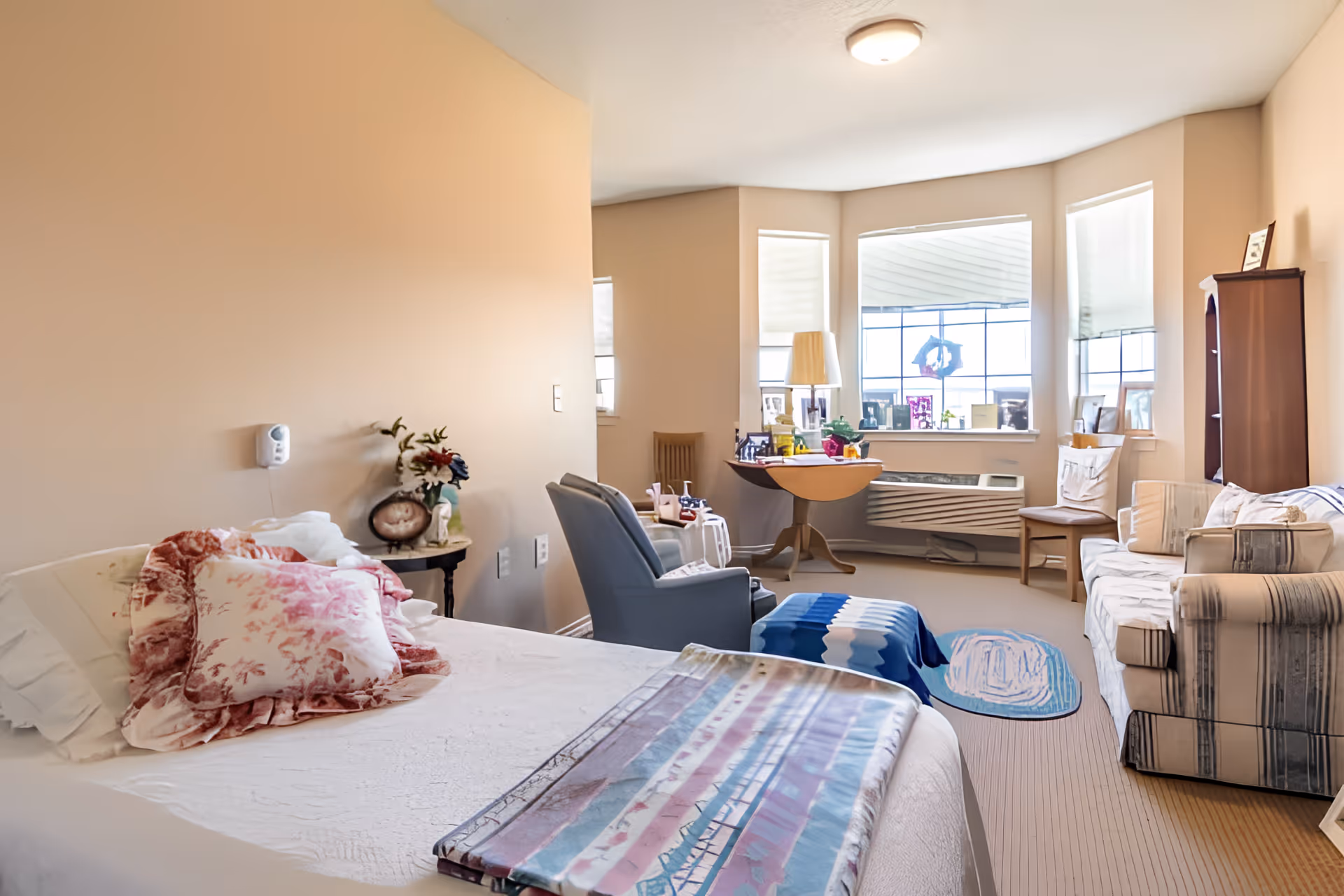 Sunny assisted-living resident room with a bed in the foreground, seating area with recliner and sofa, and a bay window with a table and decorations.