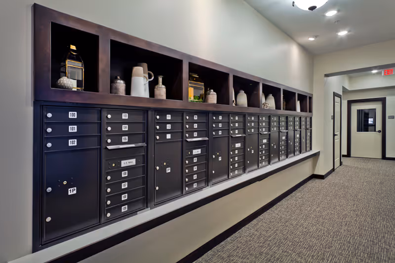 A hallway with a long row of black mailboxes mounted on the wall, each labeled with numbers. Above the mailboxes are open shelves displaying decorative items such as vases and lanterns. The hallway has carpeted flooring, light-colored walls, and recessed ceiling lights. At the end of the hallway, there is a door with a window and an illuminated exit sign above it.