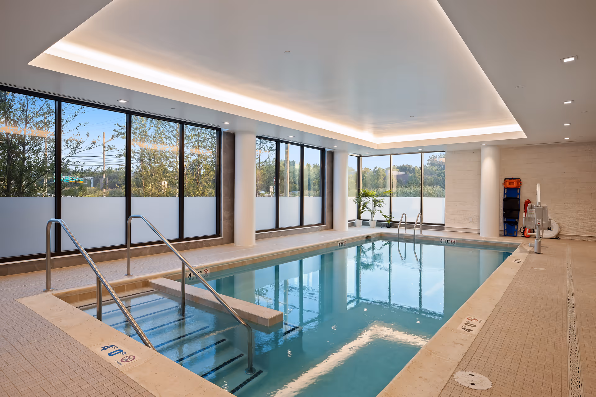 Indoor swimming pool area with large windows allowing natural light to fill the space. The pool has metal handrails and steps leading into the water. There are potted plants near the windows and safety equipment mounted on the wall. The ceiling features recessed lighting and a modern design.