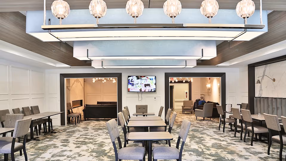 Spacious dining/common area with rows of tables and upholstered chairs, decorative overhead lighting, and a TV mounted on the far wall opening to lounge rooms.