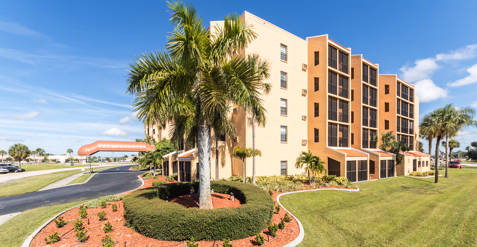 Exterior front view of a multi-story senior living building with palm trees, landscaped grounds, and a covered driveway.