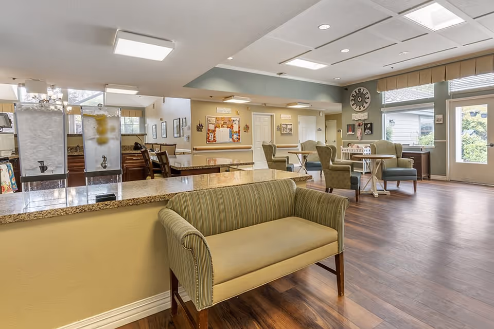 A spacious and well-lit common area in a senior living facility featuring a small striped loveseat in the foreground, a granite countertop with two beverage dispensers, several tables with green upholstered chairs, large windows letting in natural light, and a wall clock above a bulletin board.
