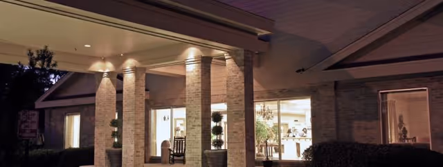 Night view of a brick senior living facility entrance with illuminated windows, columns, potted plants and a rocking chair on the covered porch.