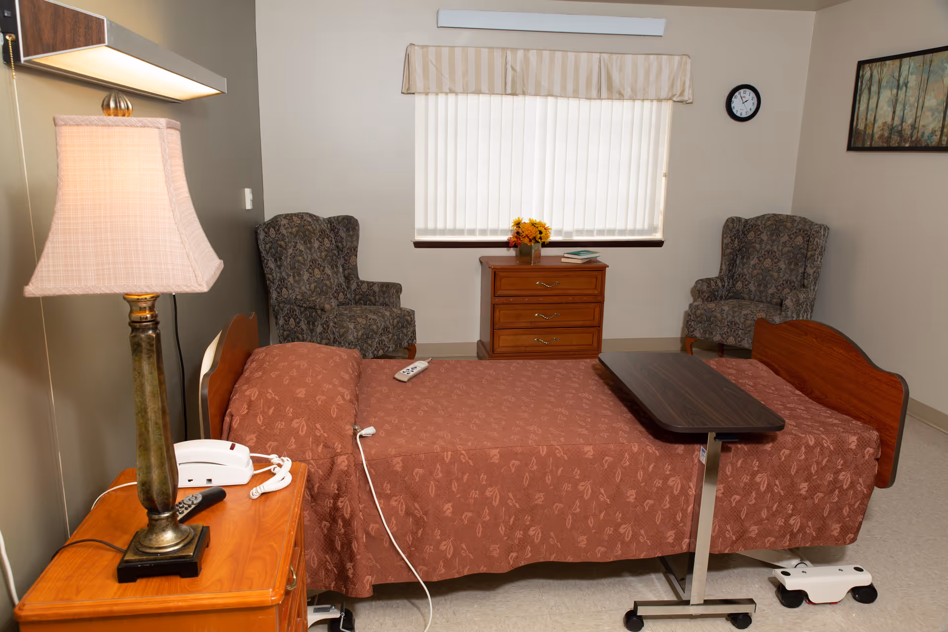 A senior living facility bedroom with a single bed covered in a reddish-brown patterned bedspread. There is a wooden bedside table with a lamp, a telephone, and a remote control. Two patterned armchairs are positioned near a window with vertical blinds and a valance. A small wooden dresser with a vase of flowers and a book sits between the chairs. A clock and a framed picture hang on the walls. A mobile overbed table is positioned over the bed.
