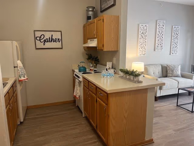 A cozy kitchen area with wooden cabinets, a white refrigerator, stove with a teal kettle, and a countertop with bottled water and a small plant. Adjacent to the kitchen is a living room with a white couch, a lamp on a side table, and decorative wall art. A framed sign on the wall reads 'Gather'.