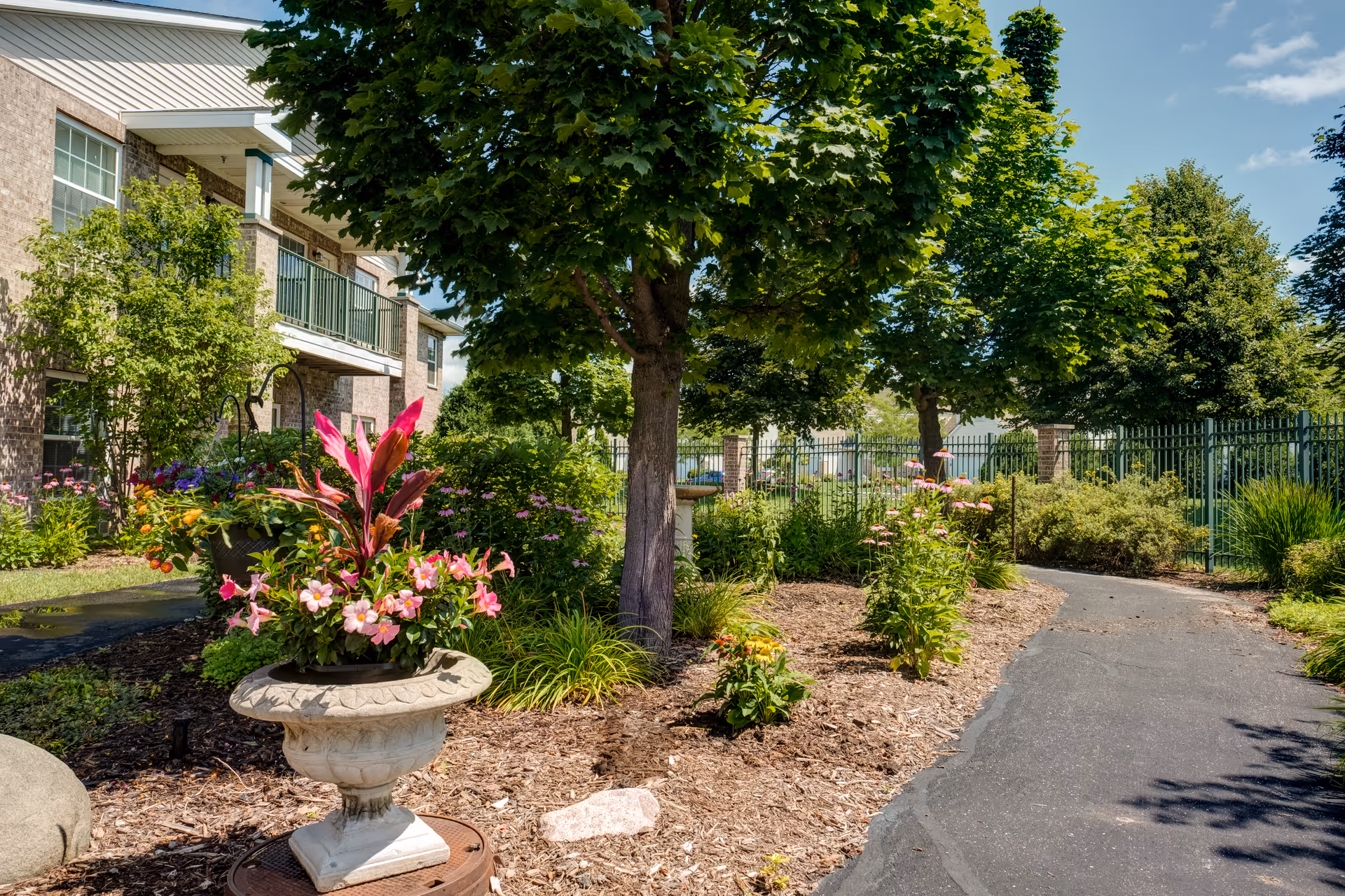 A landscaped outdoor garden area at Heritage At Deer Creek featuring a paved walking path, lush green trees, colorful flowering plants in a decorative stone planter, and a brick building with balconies in the background under a clear blue sky.